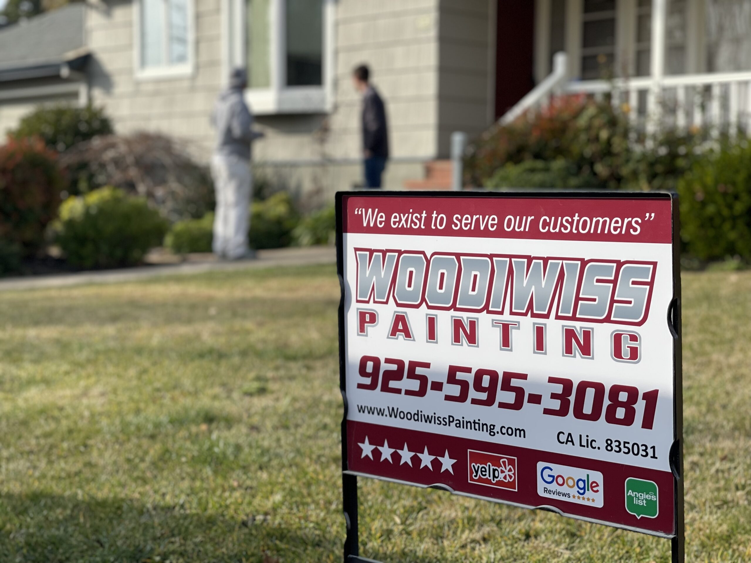 Woodiwiss Painting company sign advertising exterior house painting services A Woodiwiss Painting company sign stands on a green lawn with painters working on a house exterior.