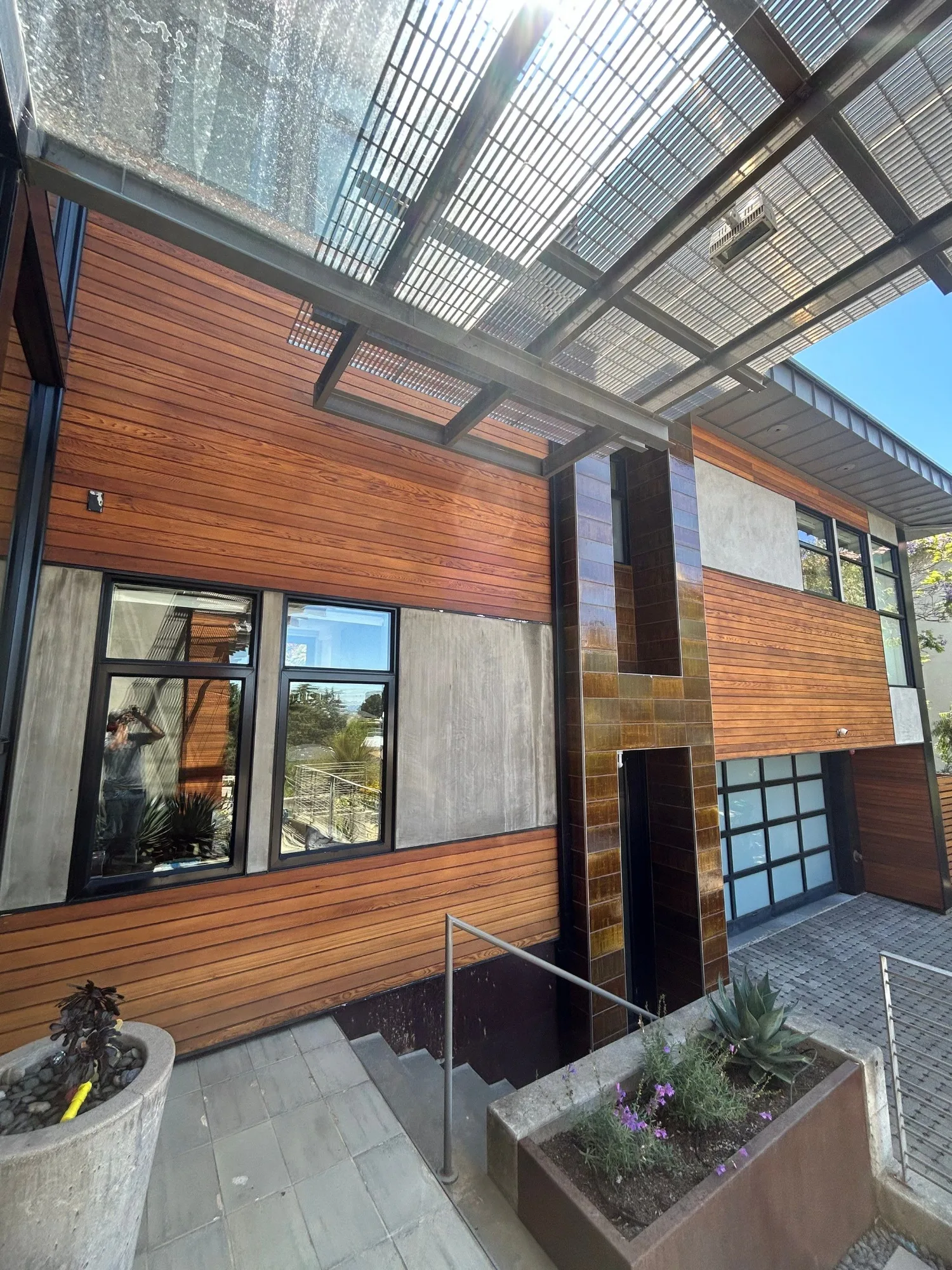 Stain After Residential Exterior Warm walnut-stained horizontal wood siding residential exterior with black-framed windows and metal pergola overhead.